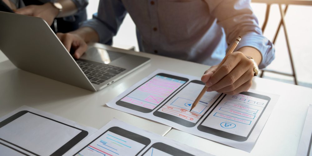 Close-up view of young male UI graphic designer planing his smartphone template while using laptop computer